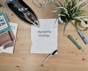 Picture of a desk with a plant, books, writing utensils, and an iron, ironing a sheet of paper with text "Marketing Strategy."