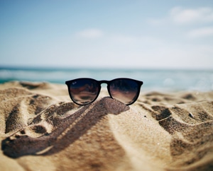 Picture of sunglasses on top of a small mound of sand with the ocean in the background.