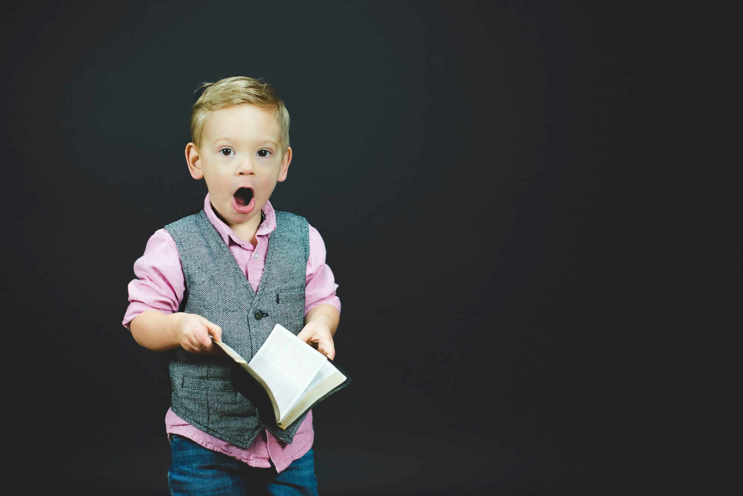 A picture of a little boy holding a Bible with his mouth open as he looks at the camera. 