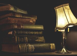 A picture of a stack of old books in a dark room with a lamp whose shade is tilted.