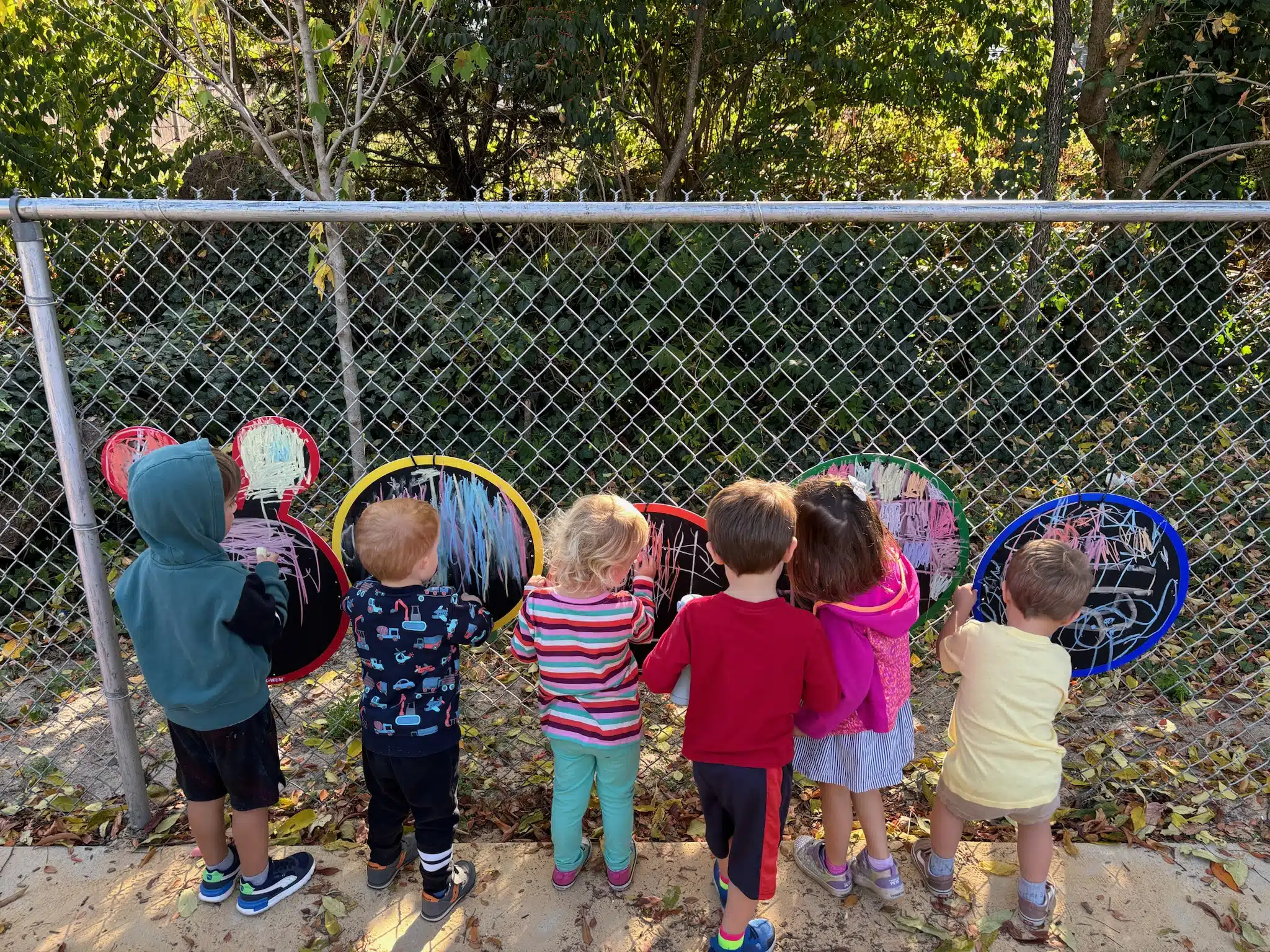 Six children drawing with chalk on colorful boards attached to a chain-link fence in an outdoor setting surrounded by trees.
