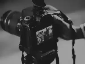 A person holds a camera displaying a blurred close-up of a group photo on its screen. Dark, monochrome ambiance.