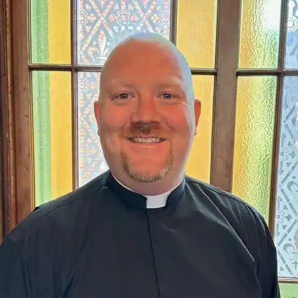 A person in clerical attire stands indoors, smiling, in front of decorative, patterned stained glass.