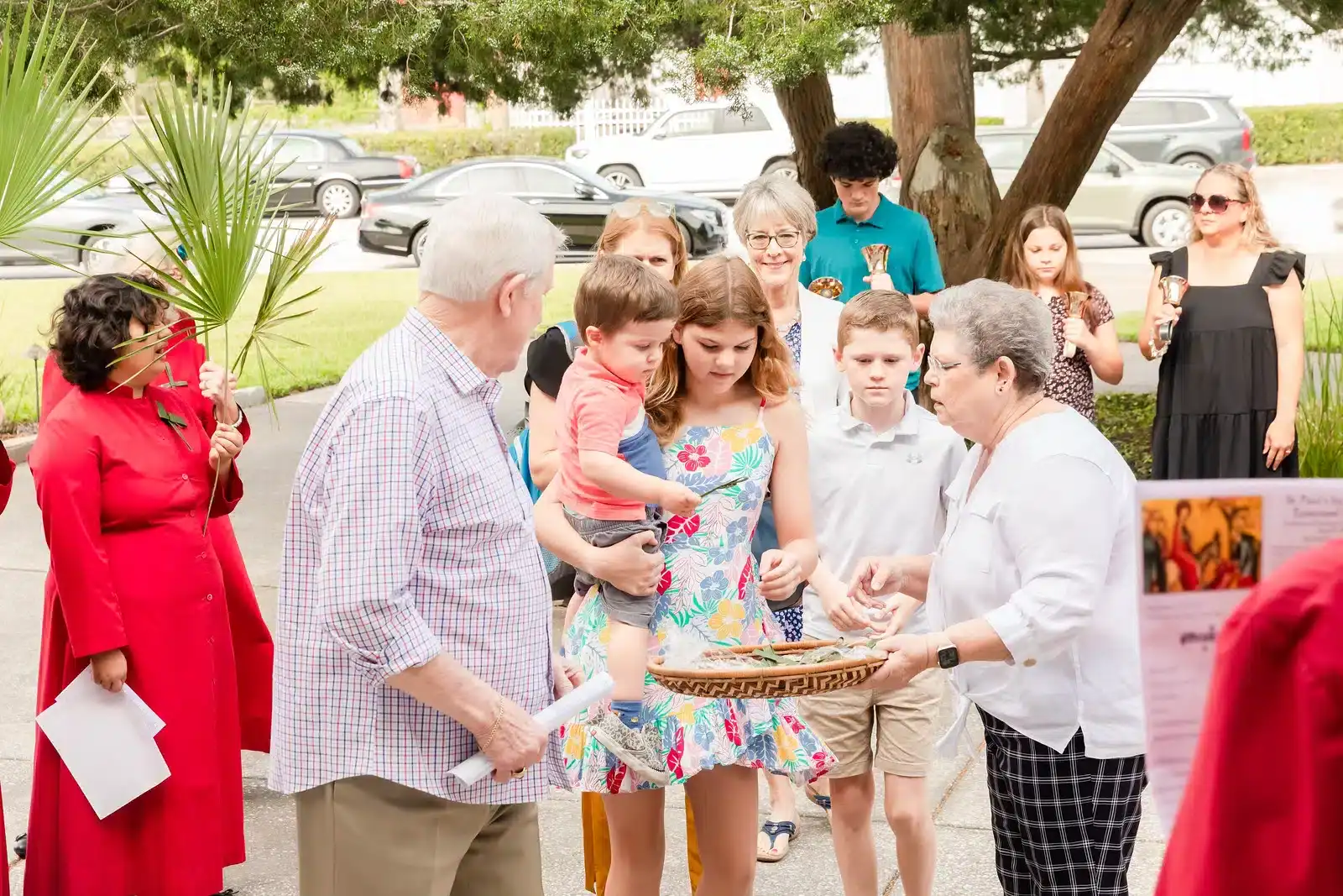 A group of people gather outside, exchanging items from a basket. Palm leaves and bells are held by participants.