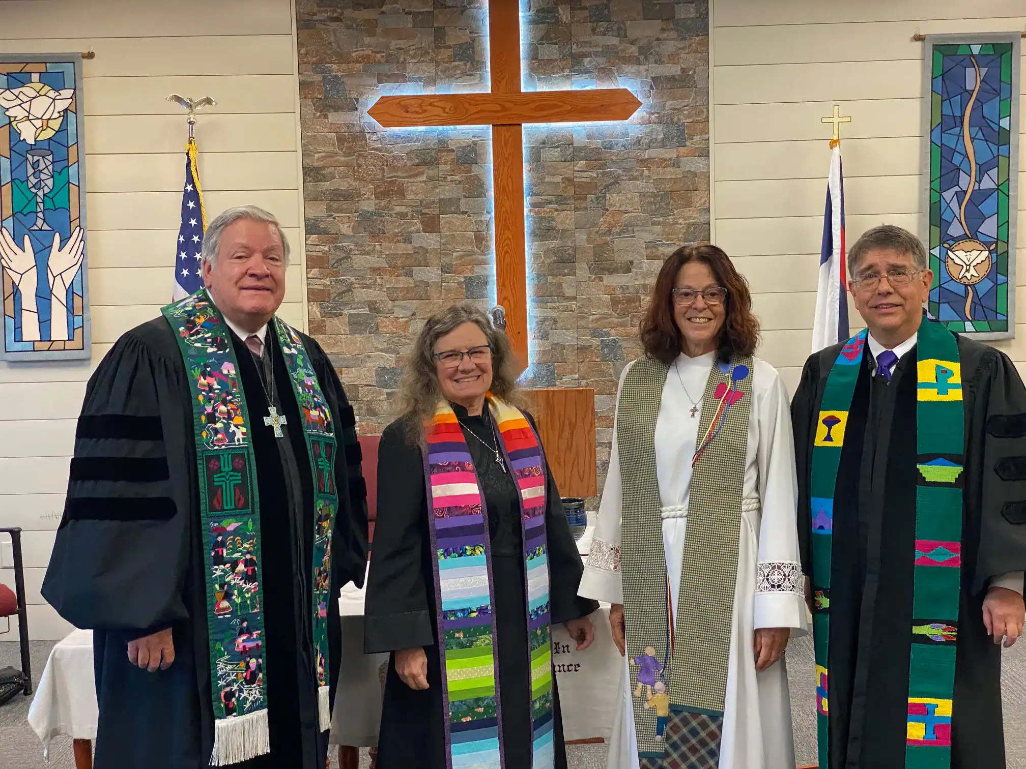 Clergy and congregation members standing in church with cross and stained glass windows.