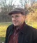 Headshot of Mike Croghan wearing a flat cap and glasses, standing outdoors in front of a grassy field with bare trees in the background