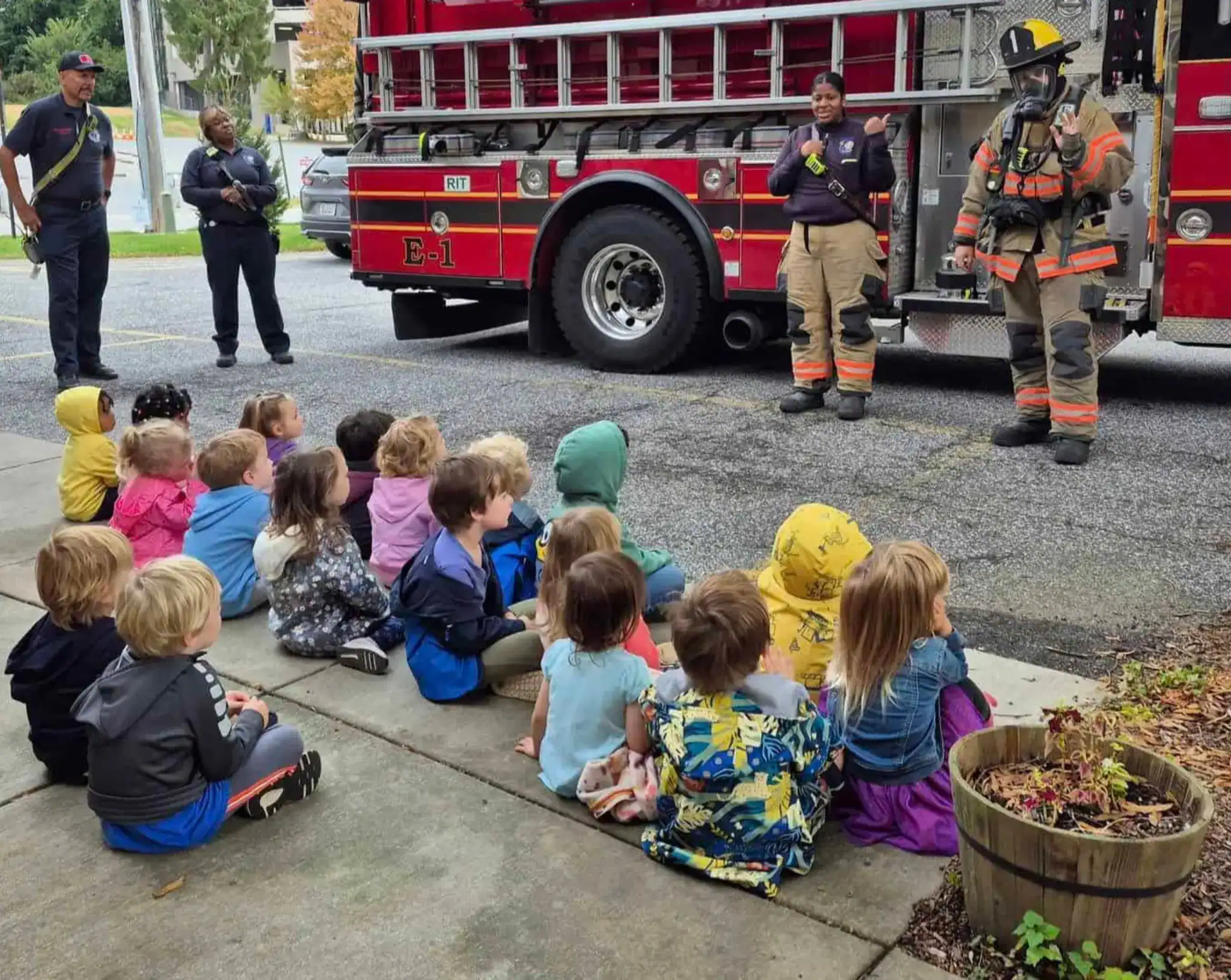 Preschool children sit on a sidewalk watching Towson firefighters and a fire engine during a safety demonstration, with one firefighter in full turnout gear and mask.